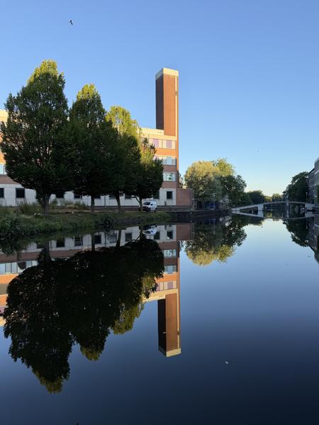 This serene photo captures a tranquil canal scene at Valckenierstraat, Weesperbuurt, Amsterdam, with a prominent modern building and its tower reflecting perfectly in the calm water